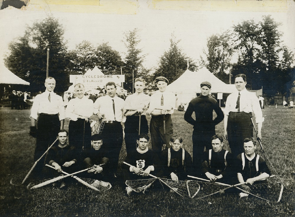 A black and white photograph of a lacrosse team posing in two rows in Town Park; front row 6 men seated on grass with lacrosse sticks crossed over legs, 7 men standing in a row behind; background park setting with three large white tents and people milling around