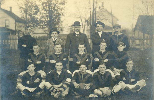 A black and white photograph of a football (soccer) team posing in three rows, five in uniform sit on grass with middle boy holding a ball, second row of 5 in uniform seated on a bench, last row of 5 standing behind; four houses and a picket fence visible in background