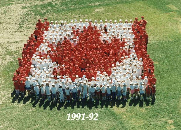 A colour photograph taken from above of a group of students dressed in red or white standing on grass in the formation of the Canadian flag