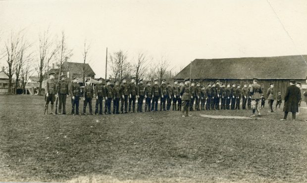 A black and white photograph of a group of young boys in uniforms with rifles, lined up in a single row; two small boys and several men, some in uniform, observe from the right side; a large rectangular wooden building and several houses visible in the background