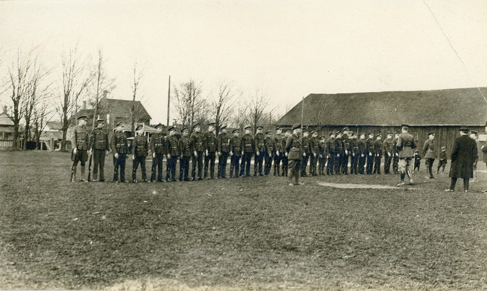 A black and white photograph of a group of young boys in uniforms with rifles, lined up in a single row; two small boys and several men, some in uniform, observe from the right side; a large rectangular wooden building and several houses visible in the background