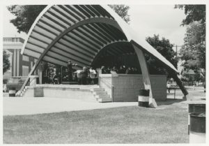 A black and white photograph of a bandshell consisting of a raised concrete platform reached via two sets of five stairs on either end at the front, the whole covered by a parabolic shaped roof supported by two curved arches; a band performs on the platform