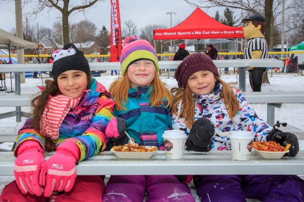 A colour photograph of three girls in brightly coloured snowsuits sitting on bleachers in a park; two girls have a container of french fries and a covered paper beverage container in front of them; an inflatable figure of a referee, several people and tents in the background