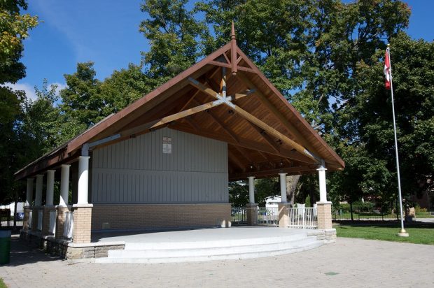 A colour photograph of a covered bandstand structure roughly square in shape with raised concrete platform, pitched wooden roof supported on ten columns along sides and central wooden and brick wall like structure; a Canadian flag on a pole stands adjacent to the front right corner; grass, trees and fencing visible in the background