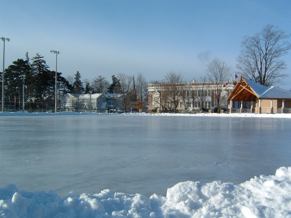 A colour photograph of a large outdoor ice rink in a park surrounded by small banks of snow; a bandstand, school building, houses and baseball diamond visible in the background