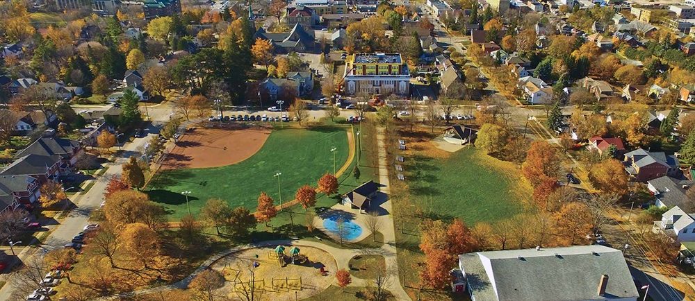 A colour image of a birds eye view of a park taken in the fall showing trees a playground and splash pad in the lower left, large rectangular white clad building with grey roof lower right, covered bandstand upper right and baseball diamond with lighting upper left; houses and buildings are visible in the background