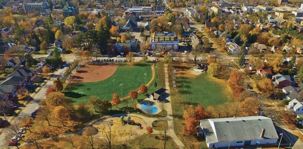 A colour image of a birds eye view of a park taken in the fall showing trees a playground and splash pad in the lower left, large rectangular white clad building with grey roof lower right, covered bandstand upper right and baseball diamond with lighting upper left; houses and buildings are visible in the background