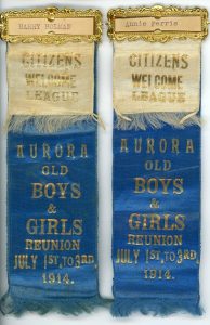 A pair of blue silk ribbons with gold lettering each bearing a secondary overlay of cream silk ribbon with gold lettering both ribbons bound together at the top with a gold rectangular frame enclosing a piece of paper with a typed name