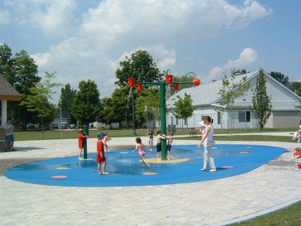 A colour photograph of seven young children and one woman carrying a baby play in splash pad consisting of a painted blue oval surface with central green pole supporting five red buckets in a spoke pattern that are being filled with water; eight painted circles on the surface of the pad feature water spouts and one a green waterpump; entire area surrounded by interlocking grey stones; several people visible along the edges; large rectangular white wooden building in the background along with trees, grass and two houses