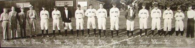 A black and white panoramic photograph of a group of 17 men standing in a straight line in a grass field, all put four men at left are wearing baseball uniforms; name of each man written in white ink appears directly beneath; trees and wooden building appear in background