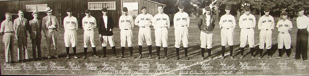 A black and white panoramic photograph of a group of 17 men standing in a straight line in a grass field, all put four men at left are wearing baseball uniforms; name of each man written in white ink appears directly beneath; trees and wooden building appear in background