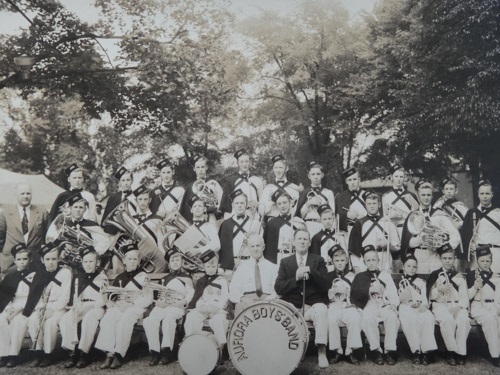 A black and white photograph of a boys band dressed in uniforms and most holding instruments arranged in three rows, front row sitting; taken in a park setting