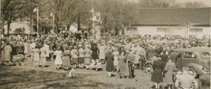 A somewhat blurry black and white photograph of a large gathering of people, most with their backs facing the camera, in a park surrounding a raised platform at the left side with four posts bearing Union Jack flags, upon which are seated about a dozen people; large white clad building and several trees and houses in the background. In the foreground there are two boys on the grass dressed in military style uniform. 