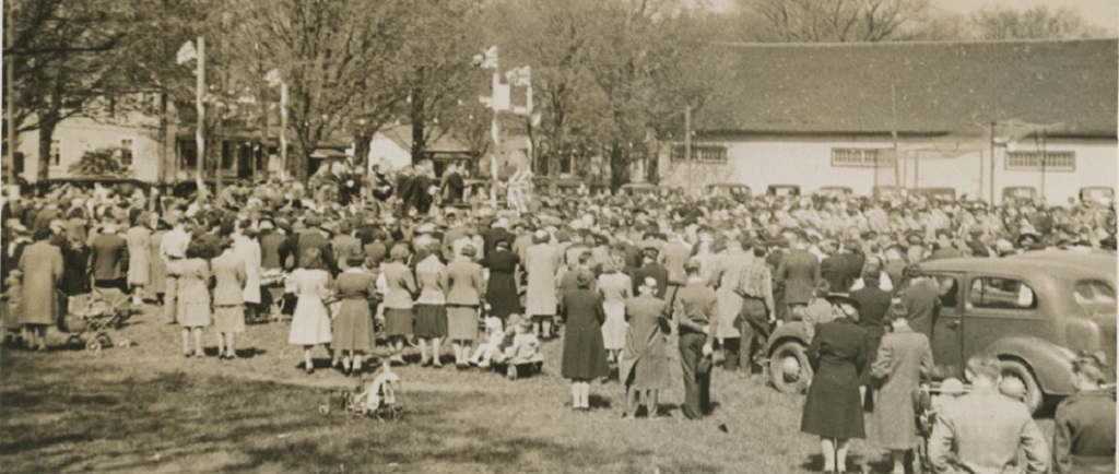 A somewhat blurry black and white photograph of a large gathering of people, most with their backs facing the camera, in a park surrounding a raised platform at the left side with four posts bearing Union Jack flags, upon which are seated about a dozen people; large white clad building and several trees and houses in the background. In the foreground there are two boys on the grass dressed in military style uniform.