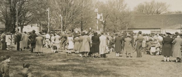 A somewhat blurry black and white photograph of a large gathering of people, most with their backs facing the camera, in a park surrounding a raised platform at the left side with four posts bearing Union Jack flags, upon which are seated about a dozen people; large white clad building and several trees and houses in the background. In the foreground there are two boys on the grass dressed in military style uniform.