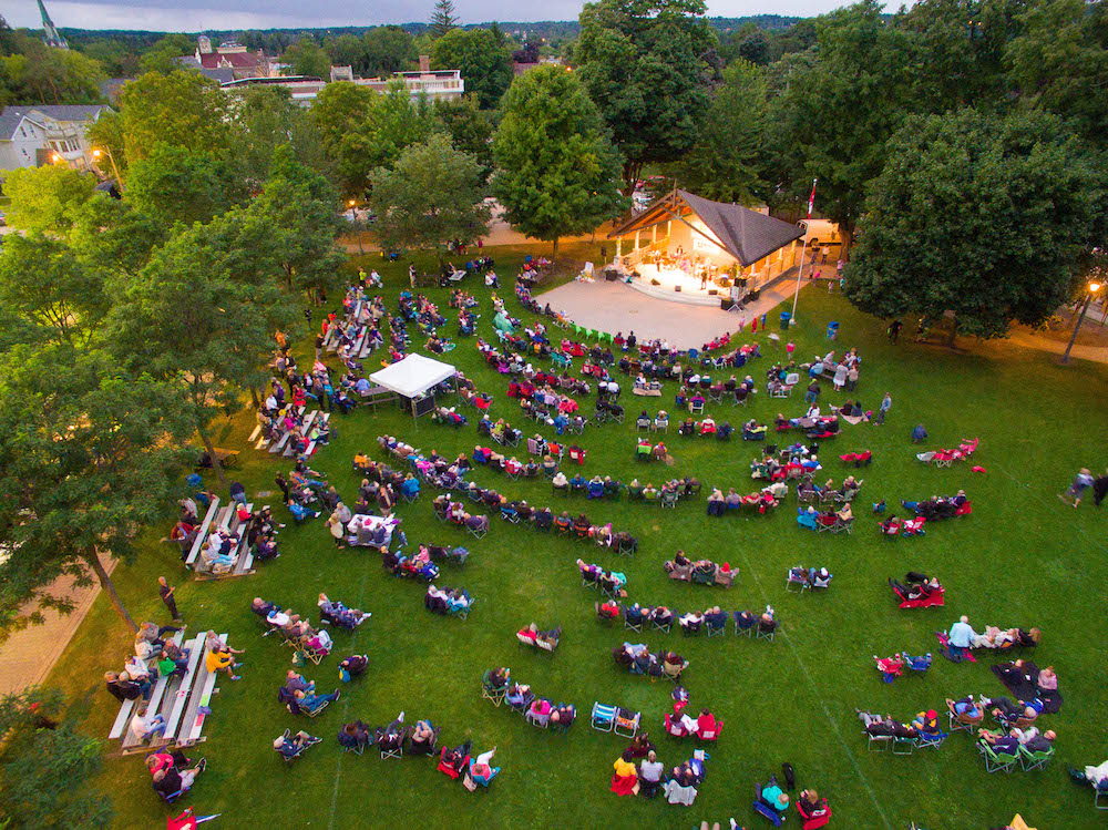 A colour photograph taken by a drone that gives a bird eye view of a section of a park that is bordered by mature trees. In the park people are seated, in no particular order, on a vast green lawn and are facing a covered performance stage that is brightly lit with musicians performing from it.