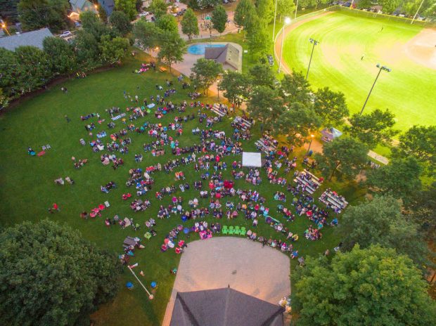 A colour image taken from a drone that gives a bird’s eye view of a large park that has a covered performance stage, splash pad and baseball diamond visible. The lawn in front of the performance stage is filled with people who are watching the stage and sitting on chairs or bleachers.