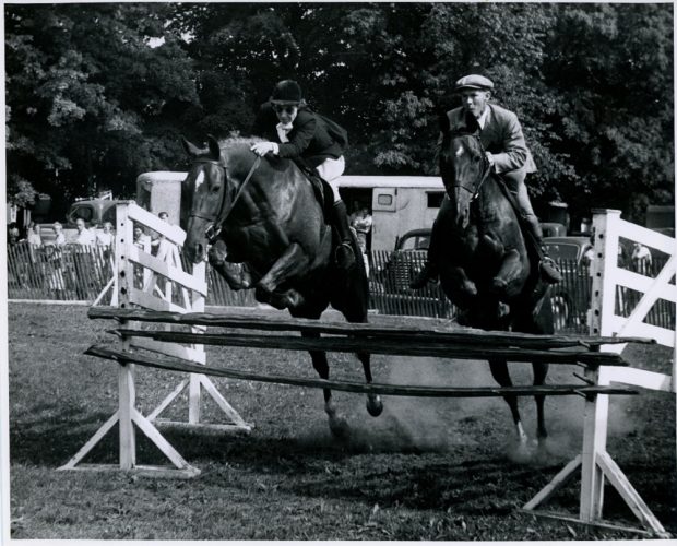 A black and white photograph of two horses and riders in a park that are simultaneously jumping over a piece of wood that is raised approximately 1 metre off the ground and is being held by two white wooden stands that are parallel to one another. The rider on the right is a man and the rider on the left is a woman. In the background people are looking on from the other side of a chest height wooden fence.