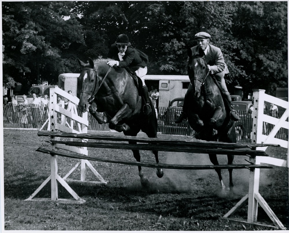 A black and white photograph of two horses and riders in a park that are simultaneously jumping over a piece of wood that is raised approximately 1 metre off the ground and is being held by two white wooden stands that are parallel to one another. The rider on the right is a man and the rider on the left is a woman. In the background people are looking on from the other side of a chest height wooden fence.