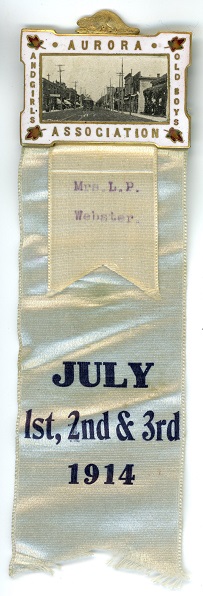 A rectangular enameled metal badge with silk ribbon attached to the bottom; the badge consists of a black and white photographic view of the main street of a town with buildings on either side set in a gold border surrounded by gold lettering and maple leaf designs on all four sides, a gold beaver design sits on the top; the silk ribbon is a light colour with blue lettering in three rows across the bottom; a smaller cream coloured ribbon sits on top of the ribbon directly underneath the badge and bears typed lettering in two rows.