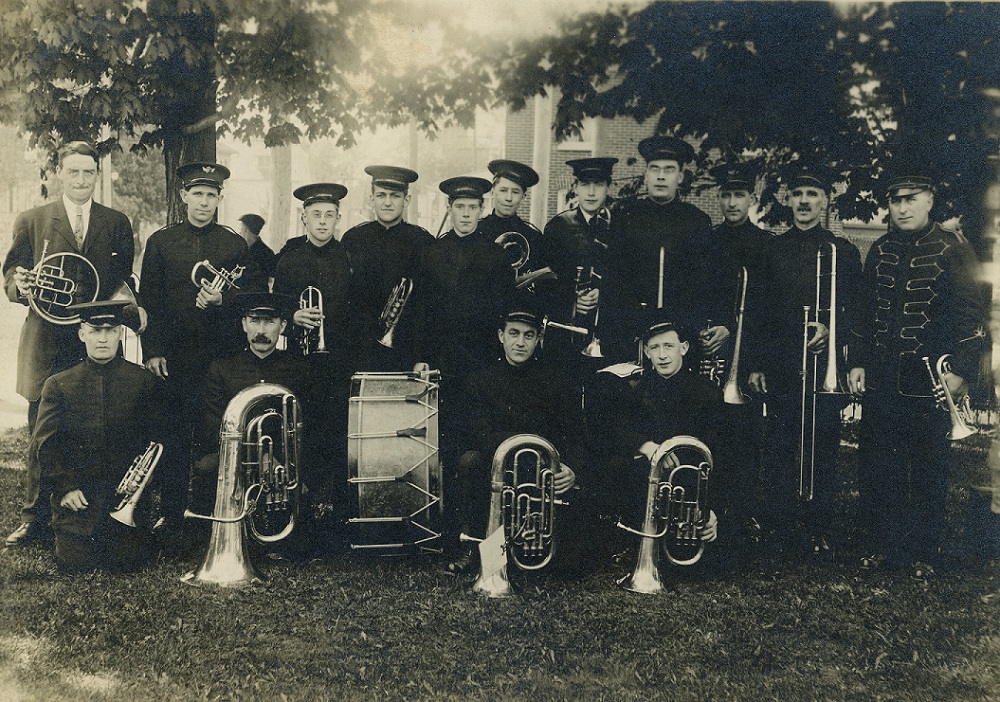 A black and white photograph of a brass band in uniforms with hats standing under trees in a park; front row kneeling; all fifteen members hold an instrument