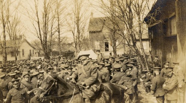 A black and white photograph of a large group of soldiers standing to the left of a wooden building; a mounted officer is in the foreground; trees and houses in the background