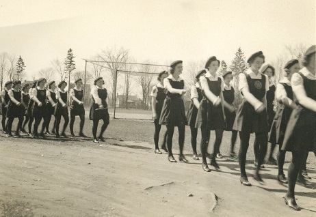 A black and white photograph depicting a line of girl cadets in uniform marching through a park; baseball diamond and houses visible in background