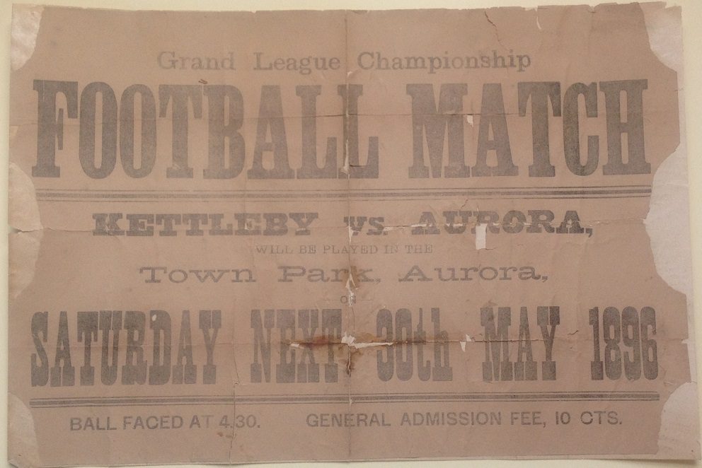 A horizontally oriented paper poster with a brown background and soft text advertising a football match in Town Park. The poster is significantly ripped along the two side edges as well as the top edge.