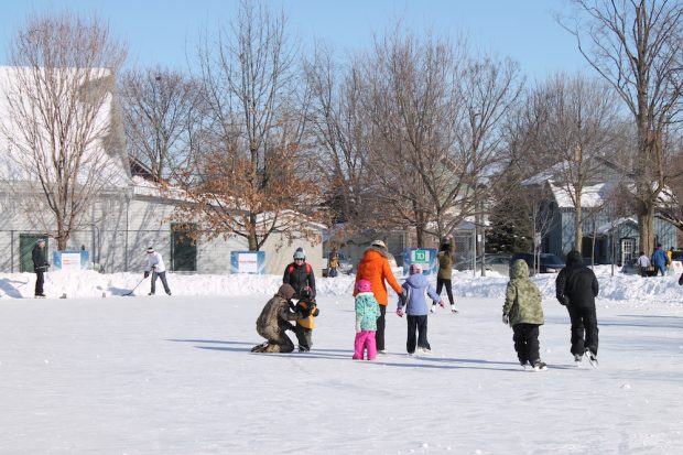 A colour image taken during the winter months that shows adults and children skating on a large outdoor rink. A couple of male skaters are holding hockey sticks. In the background to the left is a chain link fence with a large white building directly behind it, to the right two houses are visible.
