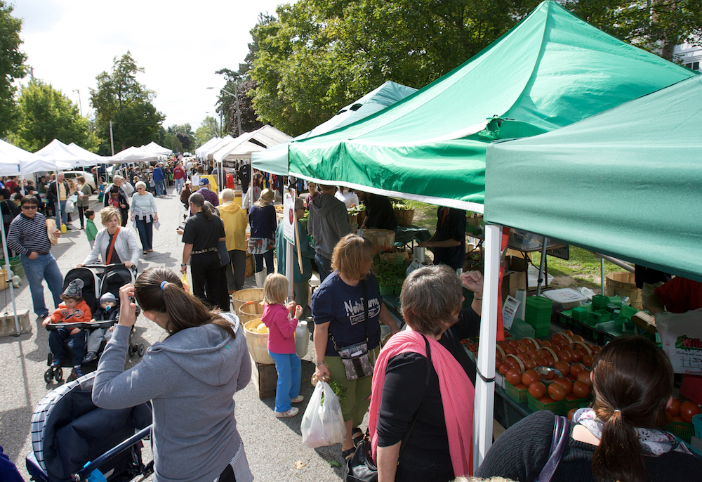 A colour photograph of a contemporary outdoor farmers’ market that has two rows of tall portable canopy tents that are running parallel to one another with a crowd of people walking between them. Underneath the tents are vendors who are selling produce and other items that aren’t clear.