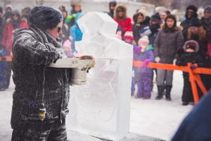 A colour image taken during the winter months showing a woman who is using a chainsaw to carve a snowman figure out a large block of ice. The background is out of focus but a crowd of adult and youth onlookers who are dressed in winter attire and gathered behind a bright orange barricade is visible.