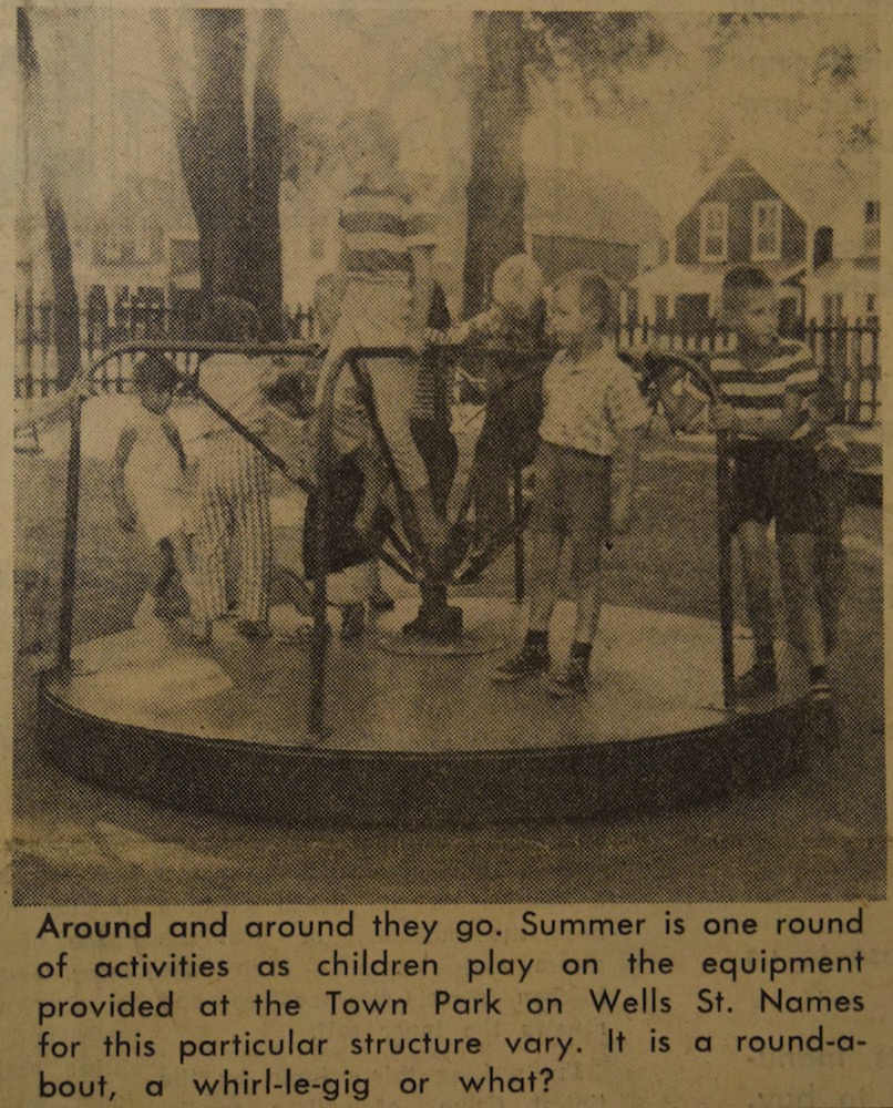 A sepia, black and grey image of nine children playing on a merry go round in an outdoor park. In the background is a picket fence and houses. There is a small caption in black text underneath the image.