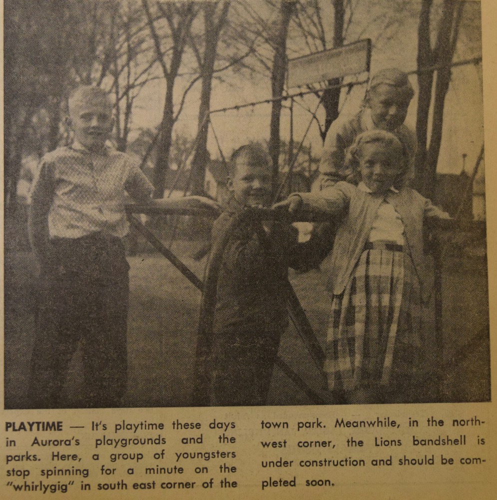 A sepia, black and grey image of three boys and one girl who are leaning on a metal rod play structure in a park. In the background a swing set and mature trees are visible. There is a small caption in black text underneath the image.