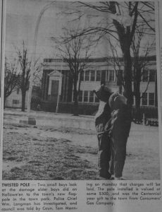 Photo en noir et blanc de deux jeunes garçons à droite dans un parc portant leur regard vers un très grand mât penché et arrivant à quelques mètres de leur tête, la base du mât sur la gauche.  À l’arrière un grand bâtiment de deux étages.  Légende en noir et blanc sous l’image.