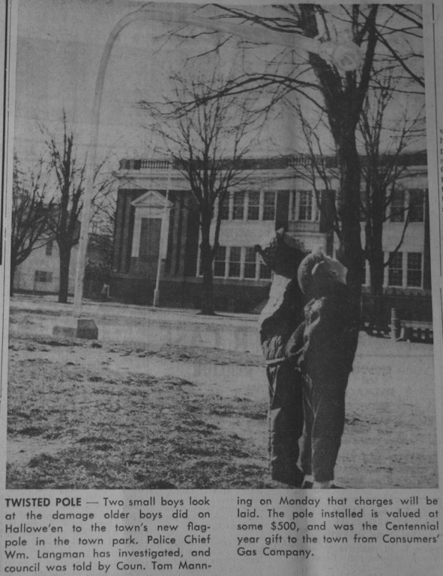 A black and white image of two young boys standing in a park. They are on the right hand side of the image and are staring up at the top of a very tall flagpole that has been bent down toward the ground and is resting a few metres above the their heads. The base of the flagpole is located on the left side, the pole bends then extends over to the right side of the image. A large two store building can be seen in the background. There is a caption of black text located directly below the image.