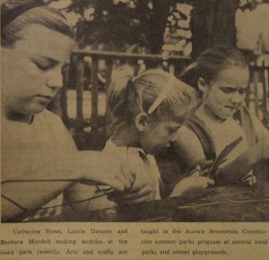 Image sépia et noir de trois filles qui utilisent une table à l’extérieur pour faire leur bricolage.  Petite légende en noir sous l’image.