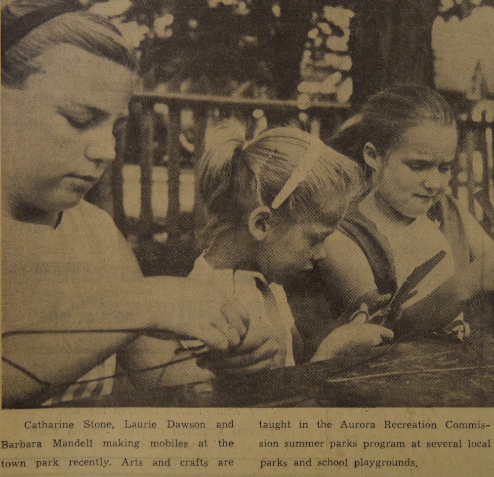 A sepia and black image of three girls who are working at an outdoor table on individual craft projects. There is a small caption in black text underneath the image.
