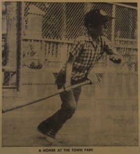 A sepia and black image of a young boy playing baseball outside. He is holding a baseball bat and appears to have just finished swinging it. The boy is staring off into the distance and looks like he is about to start running. In the background a tall chain link fence is visible as well as a shorter wooden picket fence behind it. There is a small caption in black text underneath the image.