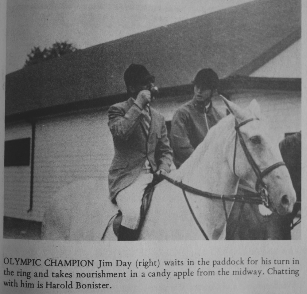 A black and white image of two men who are each sitting on top of a horse. Both men are wearing a black riding helmet and riding clothes typical of the period. The man on the left is eating a candy apple. A large building occupies the back ground and there is a caption of black text located directly below the image.