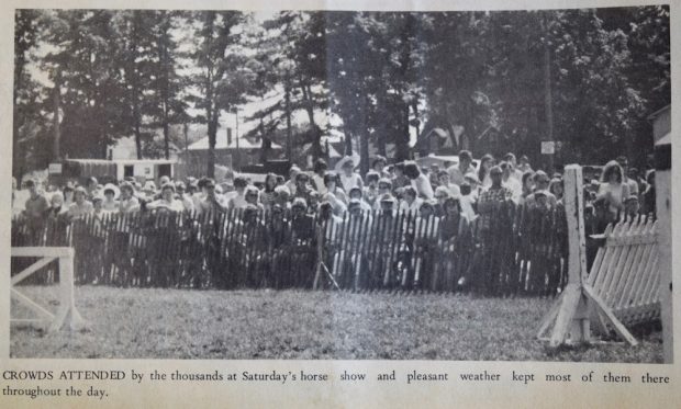 A black and white image taken in a park with a large crowd gathered in the distance behind a wooden picket fence. Behind the crowd are mature trees and houses. In the foreground pieces of white wooden structures are seen on both the left and the right side of the image. There is a caption of black text located directly below the image.