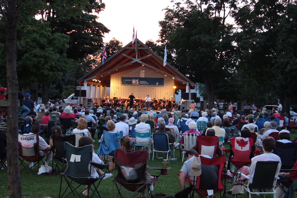 Colour photograph of a group of people seated on chairs in front of a band performing on a covered band shell in a park at dusk