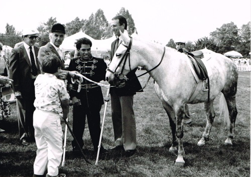 A black and white photograph of five people and a horse standing in a park setting; trees and tents visible in the background