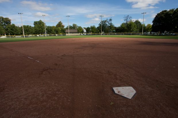 A colour image taken from behind home plate in a baseball diamond. The majority of the image shows the red dirt of the ball diamond and behind it is the grass of the outfield with a fence bordering it. In the background there is a row of mature trees and a small building close to the center of the image.