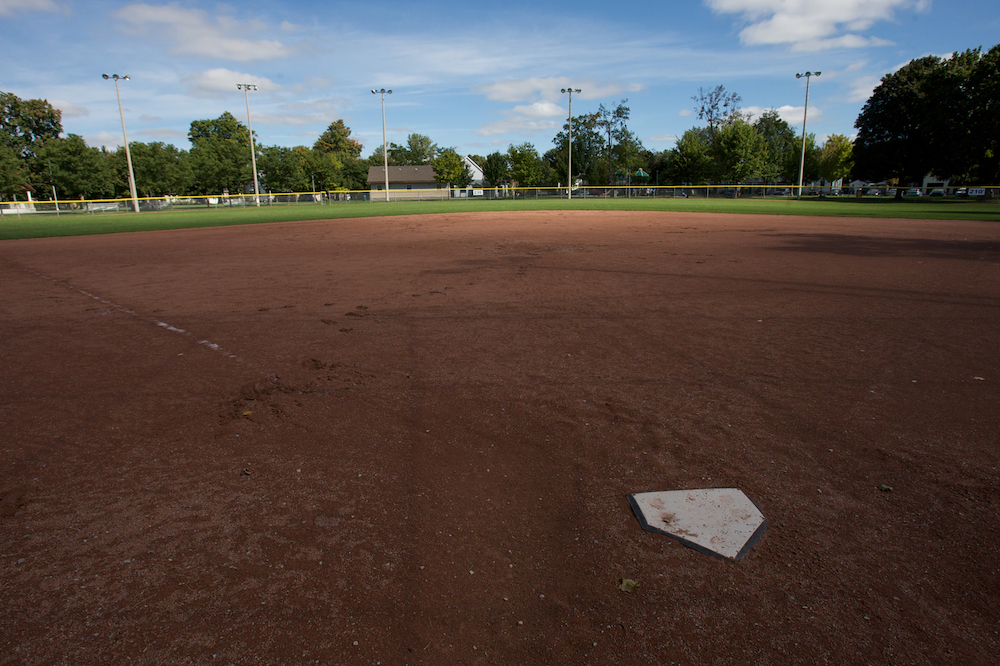 A colour image taken from behind home plate in a baseball diamond. The majority of the image shows the red dirt of the ball diamond and behind it is the grass of the outfield with a fence bordering it. In the background there is a row of mature trees and a small building close to the center of the image.