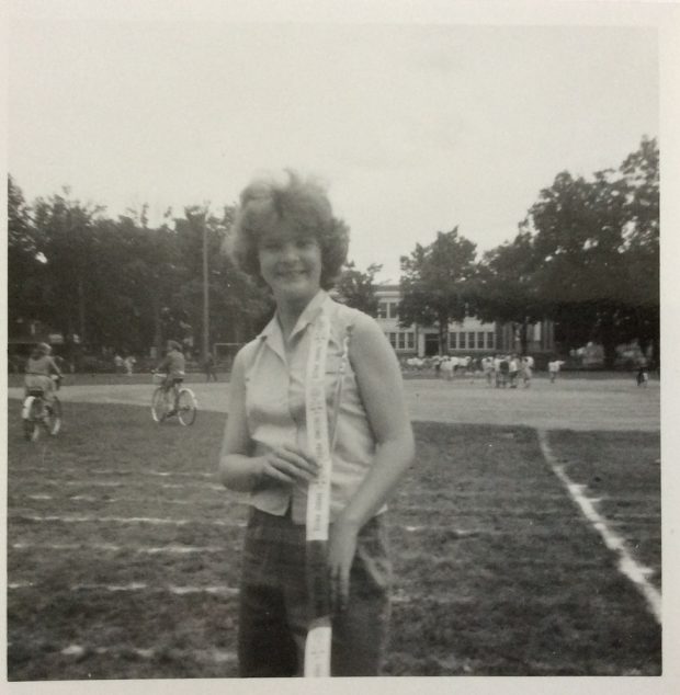Black and white image of a girl standing in a large field with chalk lines on the grass. She is in the centre of the photograph and has a banner of track and field ribbons draped over her right shoulder. In the background on the left side there is a man and a woman each riding a bike across the field, on the right side is a group of kids playing in a crowd. A two-storey school house is also visiable in the background surrounded by mature trees.