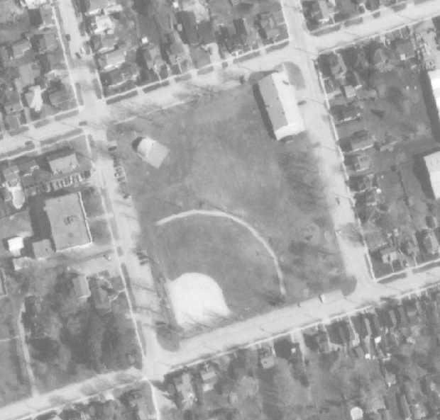 A black and white aerial photograph of Town park showing a ball diamond, band shell and Armoury building; adjacent streets and houses also visible in shot