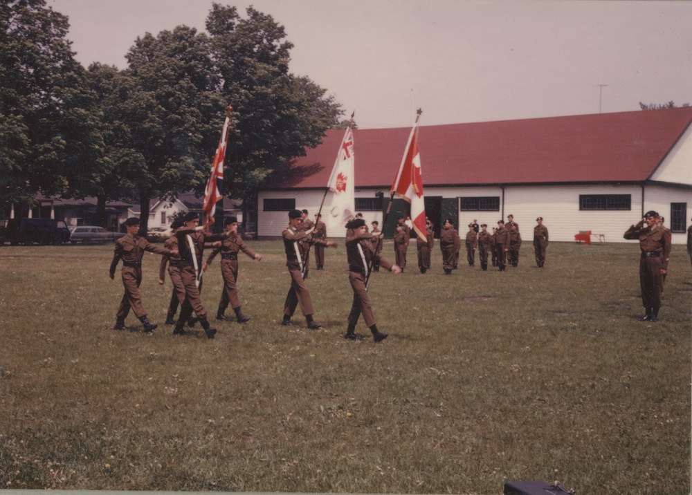 A colour photograph depicting uniformed boys parading with flags in a park setting; trees and a rectangular white clad building with red roof in the background