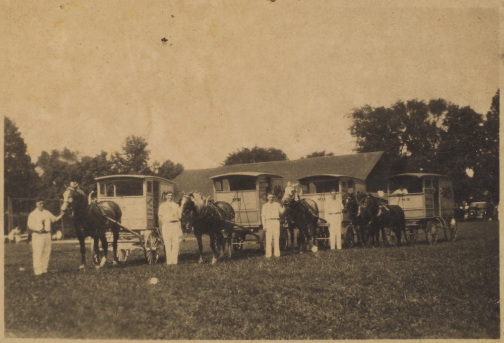 A sepia coloured image from c. 1930s showing four men dressed all in white standing in a park. To the right of each of them is a horse attached to a buggy. The man on the left side appears to be holding something in his hand. There is a fifth man seated inside of the buggy on the far right. In the background, a large roof and tall baseball diamond fence is visible.