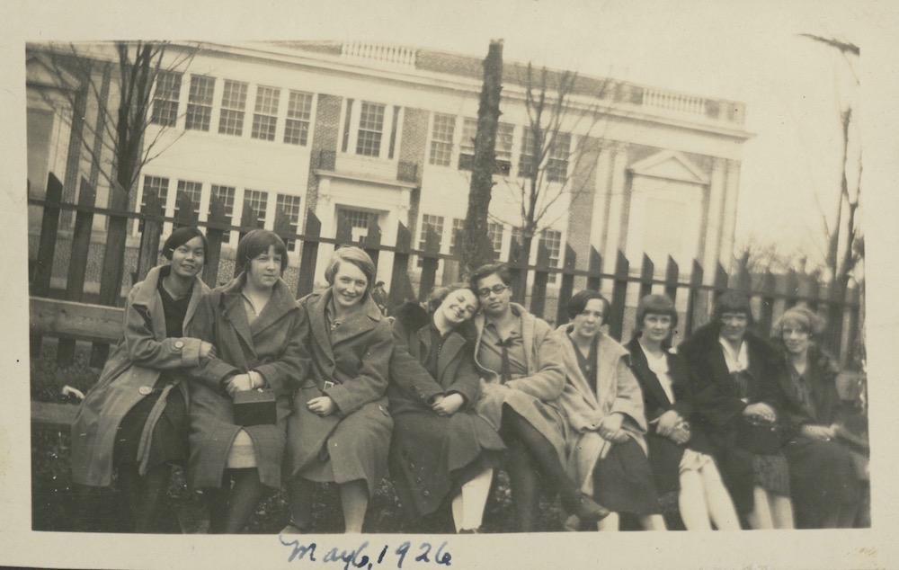 a sepia coloured photograph from 1926 showing nine girls seated on a bench with a wooden fence immediately behind them. A large two storey structure takes up the majority of the background. The image was taken on an angle so the fence and building appear to be slanted towards the right side.