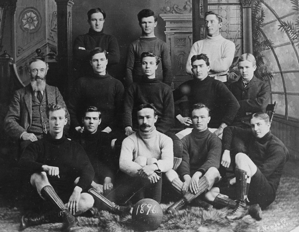 Formal black and white portrait taken inside of a photo studio featuring a football (soccer) team with three rows of men: the back row has three men standing, the middle row has five men seated on chairs, and the bottom row has five men seated on the floor. In front of the bottom row is a round ball that has 1896 written on it.